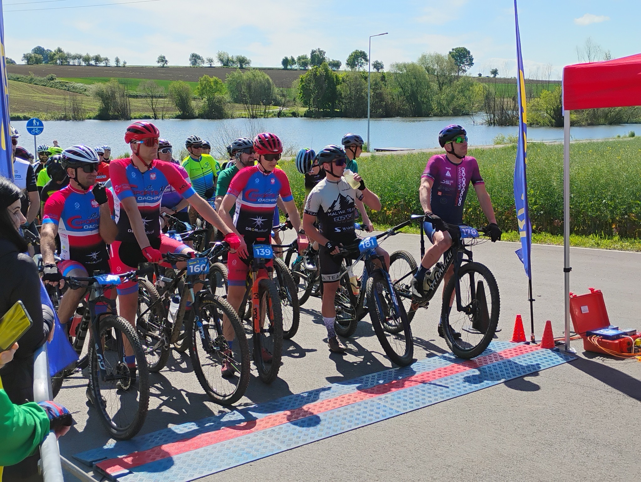 Grupa kolarzy oczekująca na&nbsp;start wyścigu, w&nbsp;tle Źr&oacute;dlana Oaza