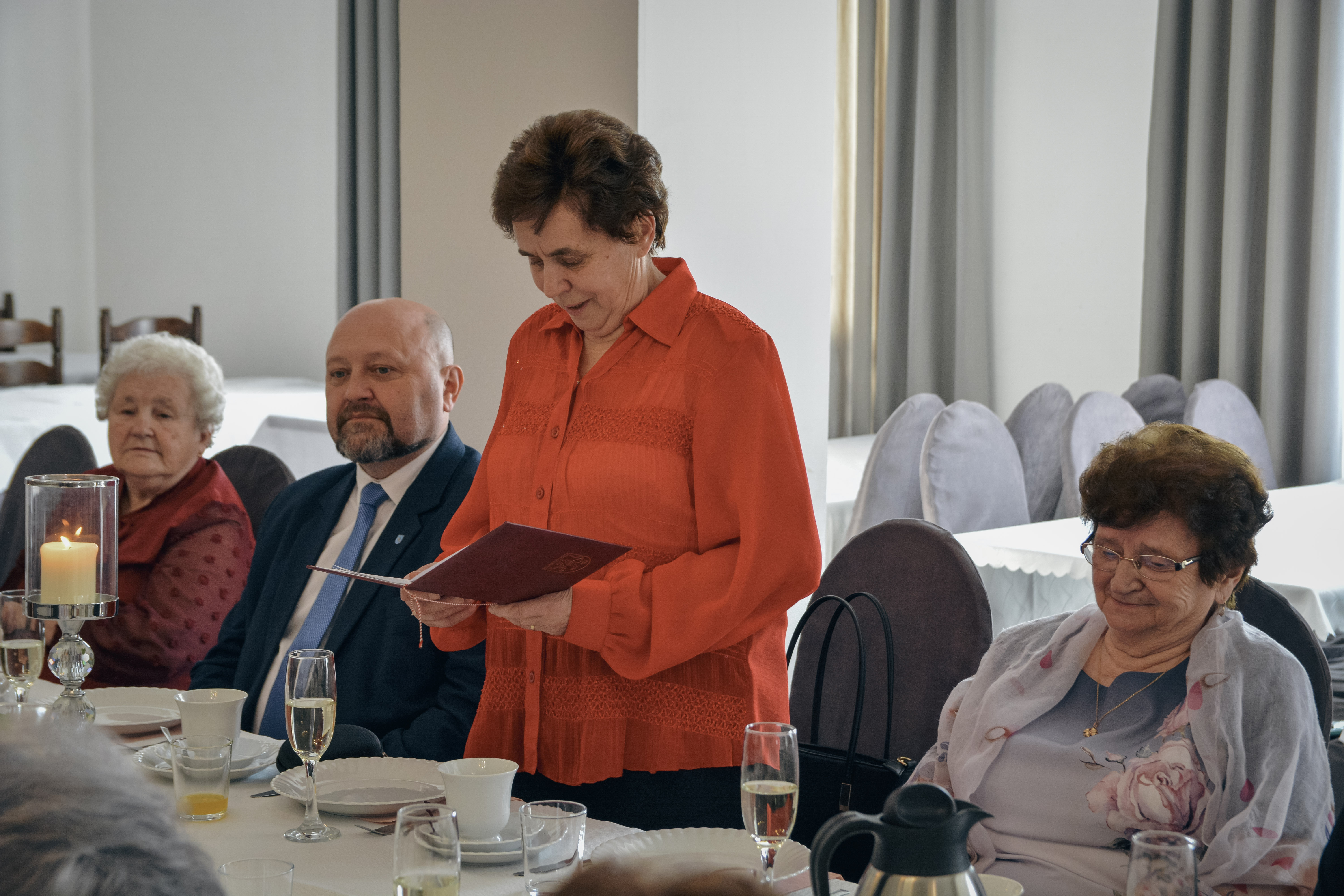 Pani w&nbsp;czerwonej bluzce stoi za stołem i&nbsp;odczytuje coś z&nbsp;kartki, obok niej przy stole siedzą mężczyzna i&nbsp;dwie kobiety