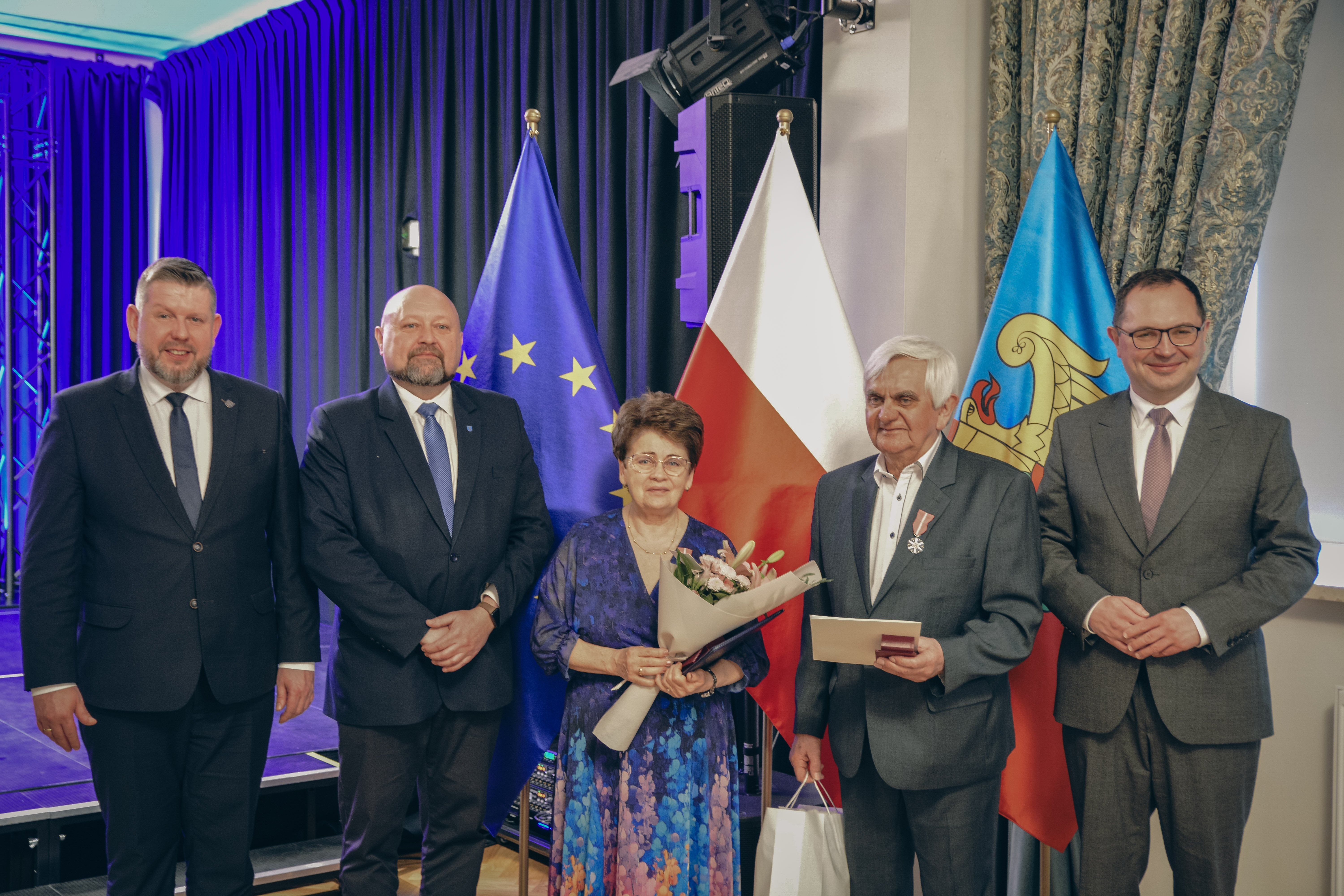 Na sali na&nbsp;tle flagi Unii Europejskiej, flagi Polski i&nbsp;flagi Wodzisławia Śląskiego stoi para obchodząca Złote Gody, obok W&oacute;jt Marklowic, Prezydent Wodzisławia Śląskiego oraz&nbsp;Wicewojewoda Śląski