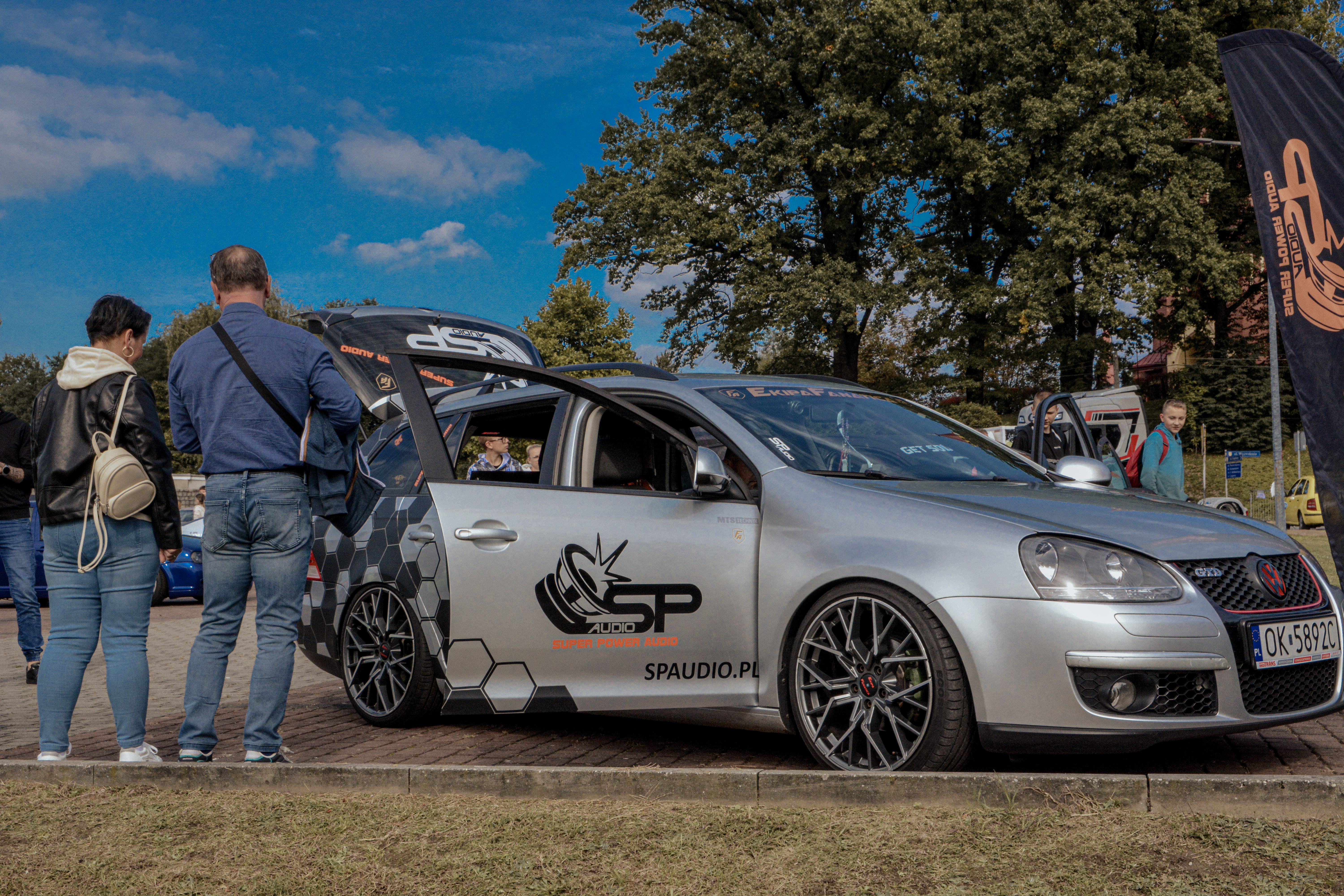 Srebrne, sportowe auto stoi na&nbsp;parkingu, przy nim stoją osoby i&nbsp;patrzą na&nbsp;samoch&oacute;d