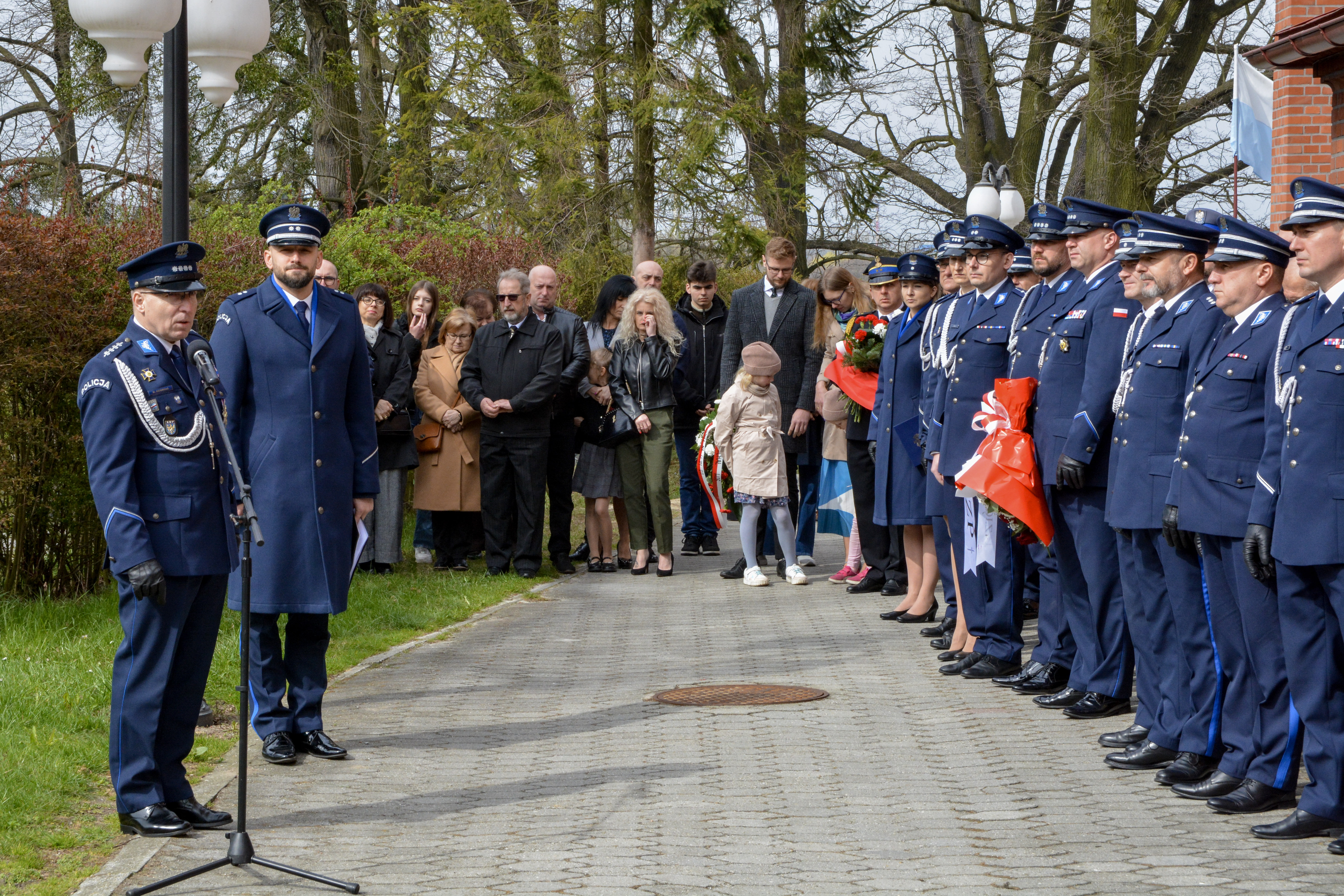 Obok budynku kościoła stoją przedstawiciele policji w&nbsp;mundurach, niekt&oacute;e osoby mają kwiaty, jeden z&nbsp;nich przemawia do zebranych przez mikrofon
