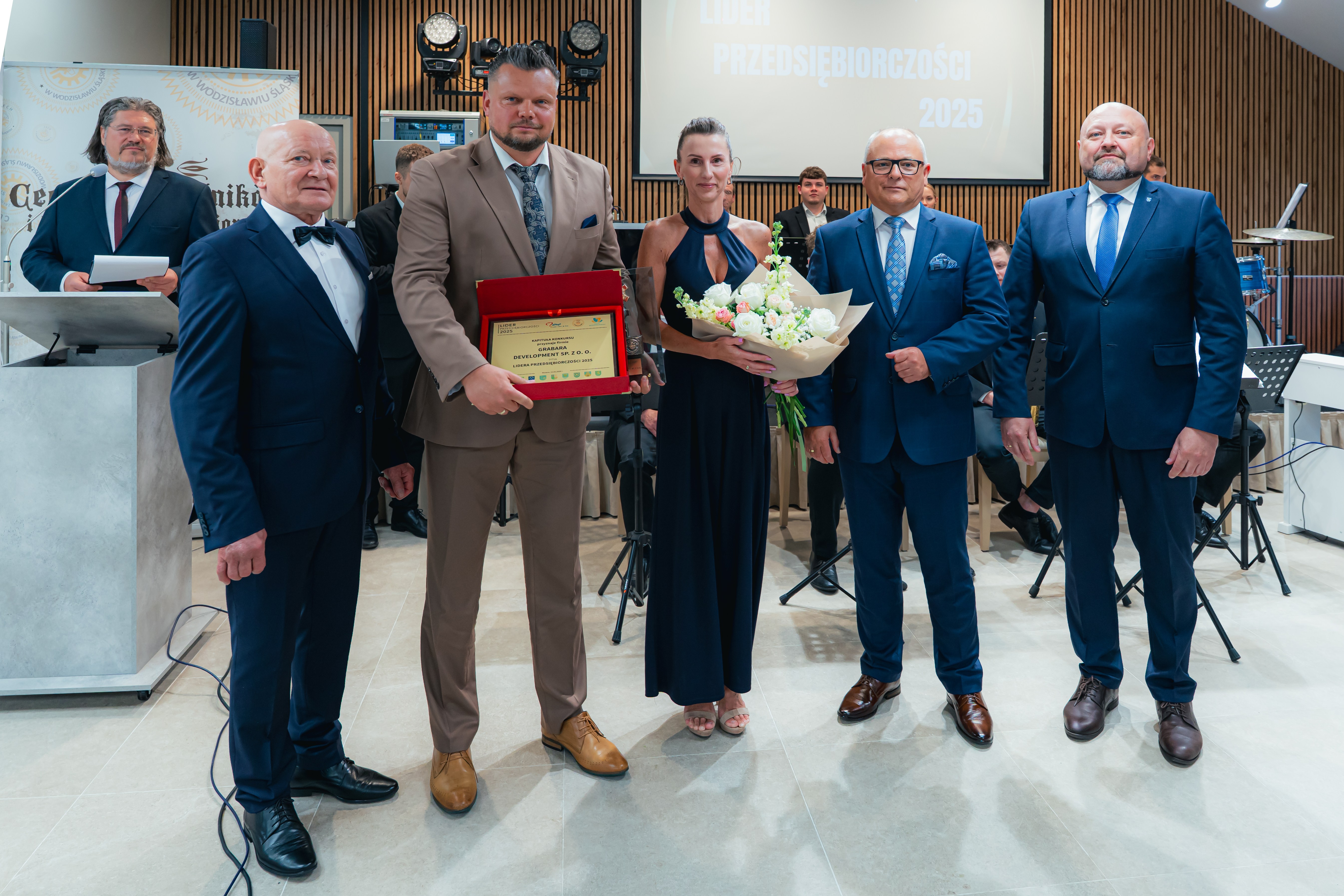 W pomieszczeniu do zdjecia pozuje kilka os&oacute;b, jeden mężczyzna trzyma pamiątkową tabliczkę w&nbsp;wyr&oacute;żnieniem dla firmy GRABARA DEVELOPMENT, kobieta trzyma bukiet kwiat&oacute;w, obok władze powiatu i&nbsp;Gminy oraz&nbsp;prezes Cechu Rzemieślnik&oacute;w
