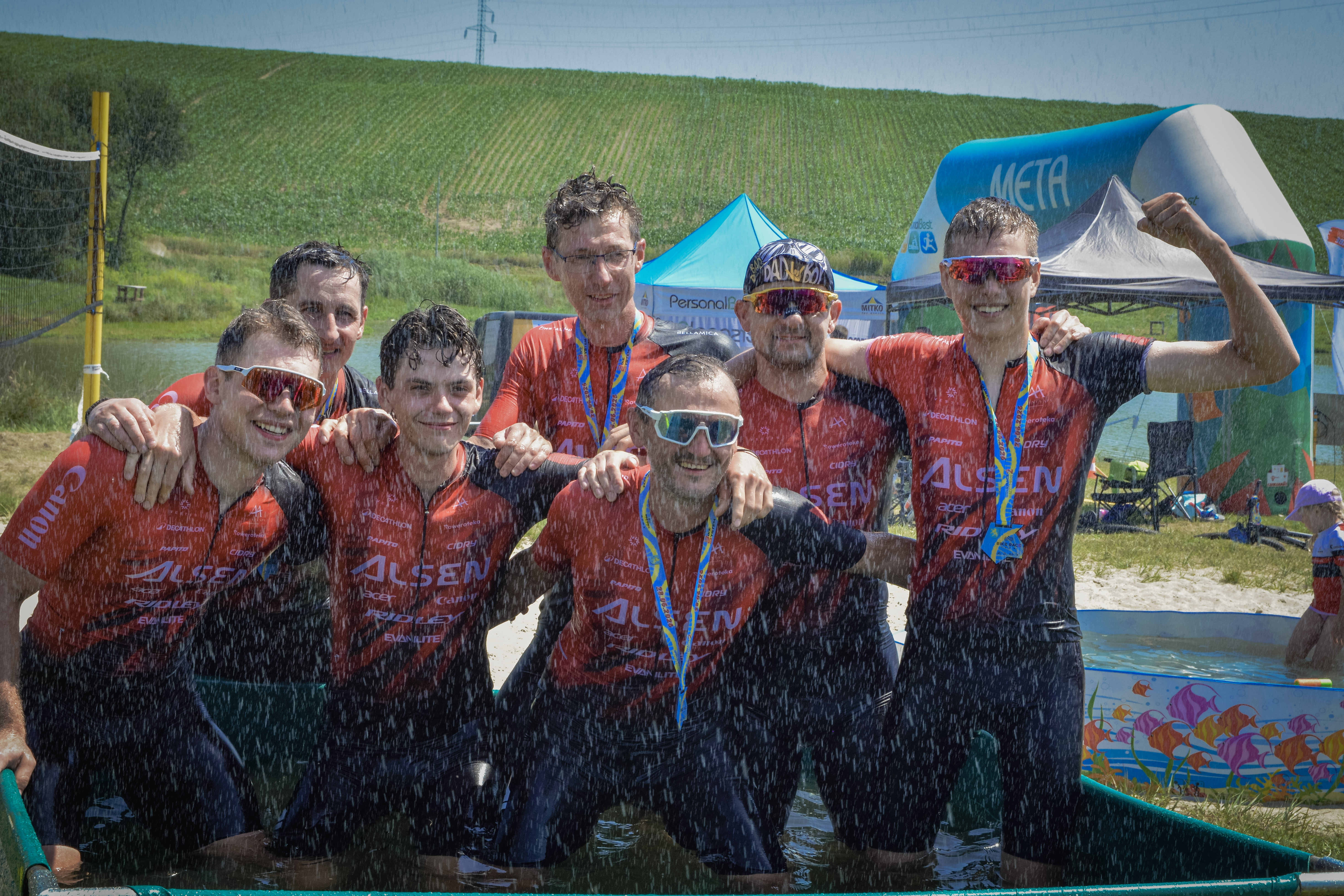7 mężczyzn w&nbsp;czerwono-czarnych strojach sportowych pozuje do zdjecia w&nbsp;basenie z&nbsp;wodą podczas wydarzenia Bike Race & Szanty