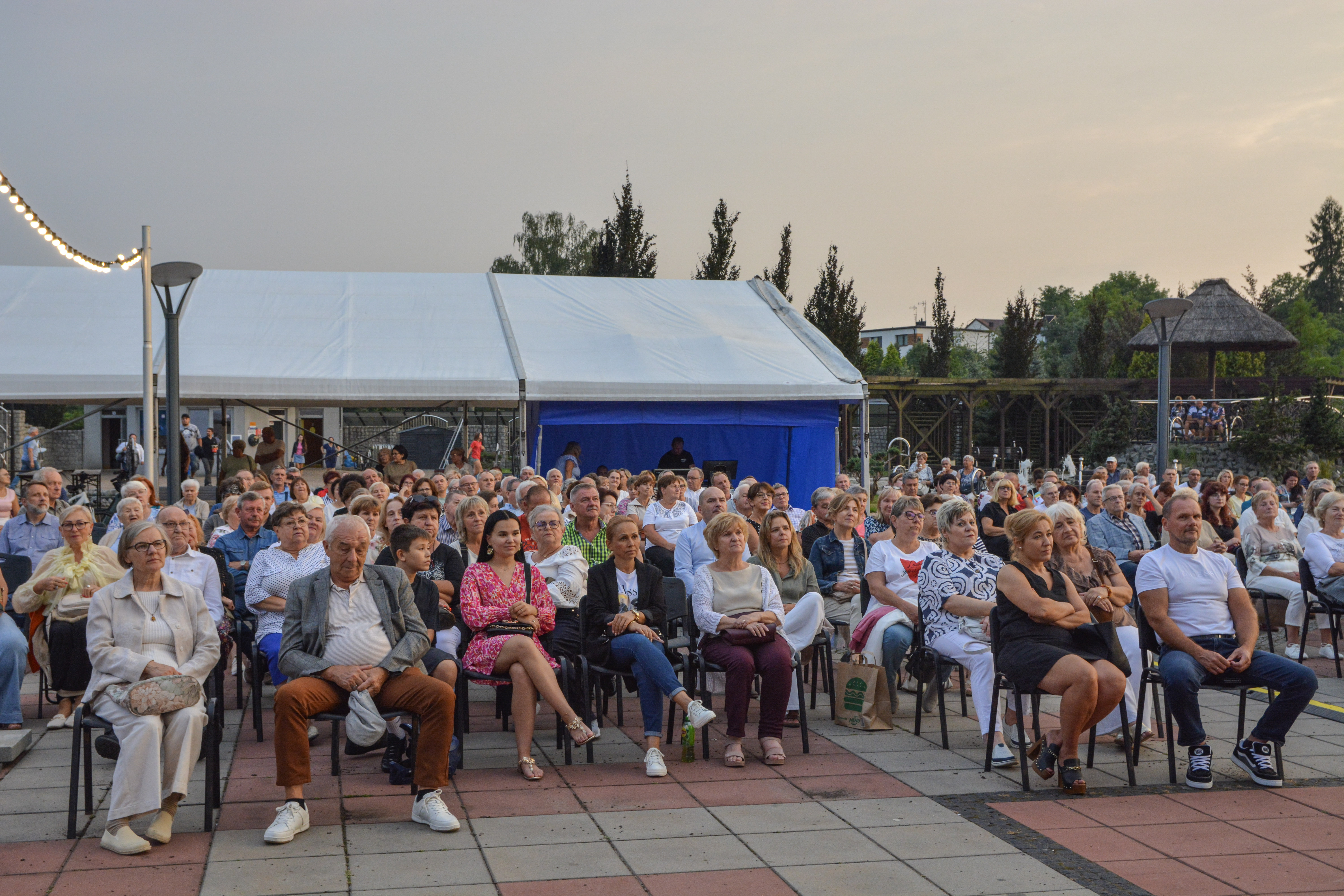 Duża grupa ludzi siedzi na&nbsp;krzesłach na&nbsp;dużym placu, w&nbsp;tle namiot eventowy, obok świetlna girlanda zawieszona na&nbsp;słupku