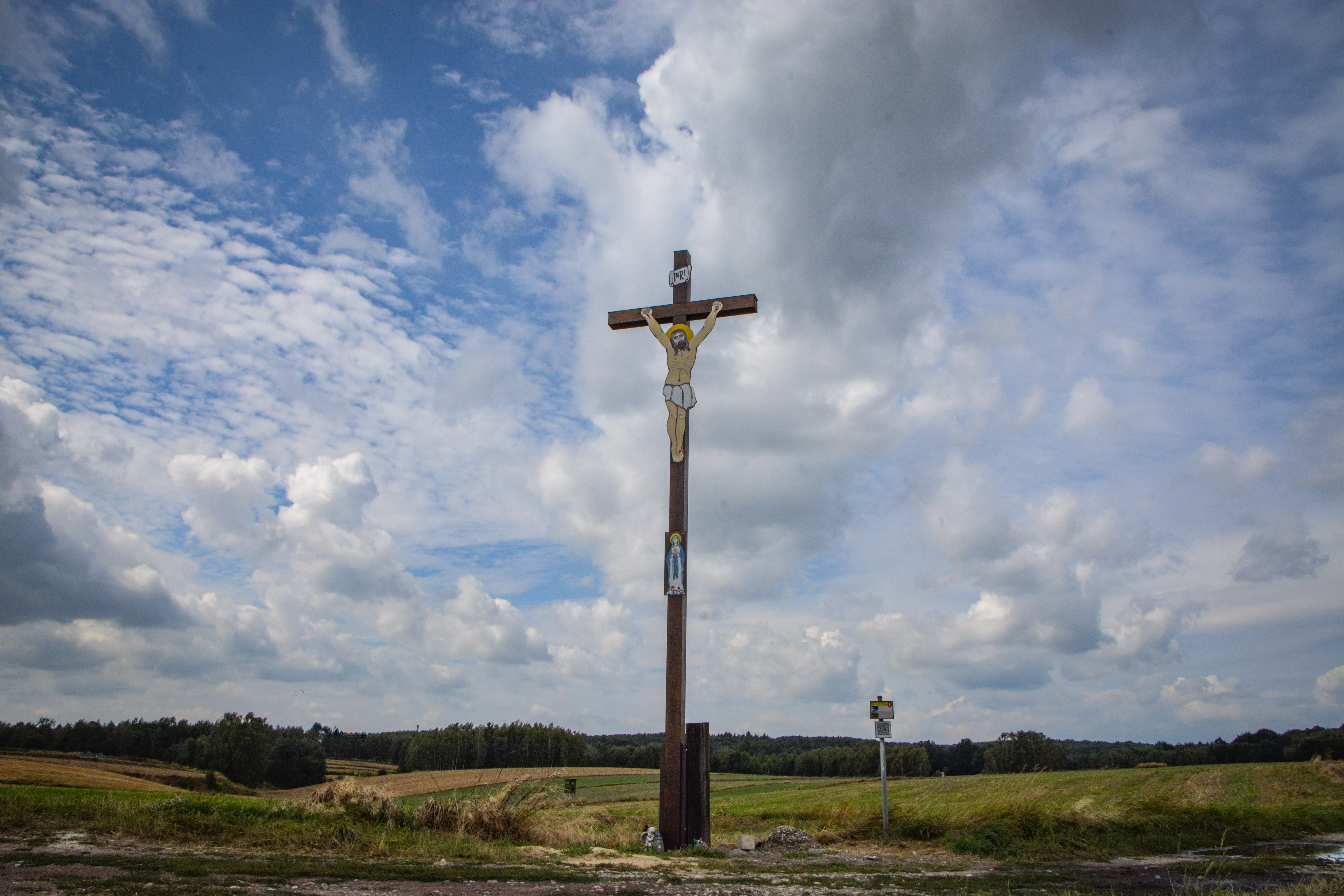 Polne rozdroże, przy nim stoi duży drewniany krzyż z&nbsp;wizerunkiem umęczonego Jezusa, pod spodem mniejszy wizerunek Matki Boskiej, w&nbsp;tle pola uprawne i&nbsp;lasy