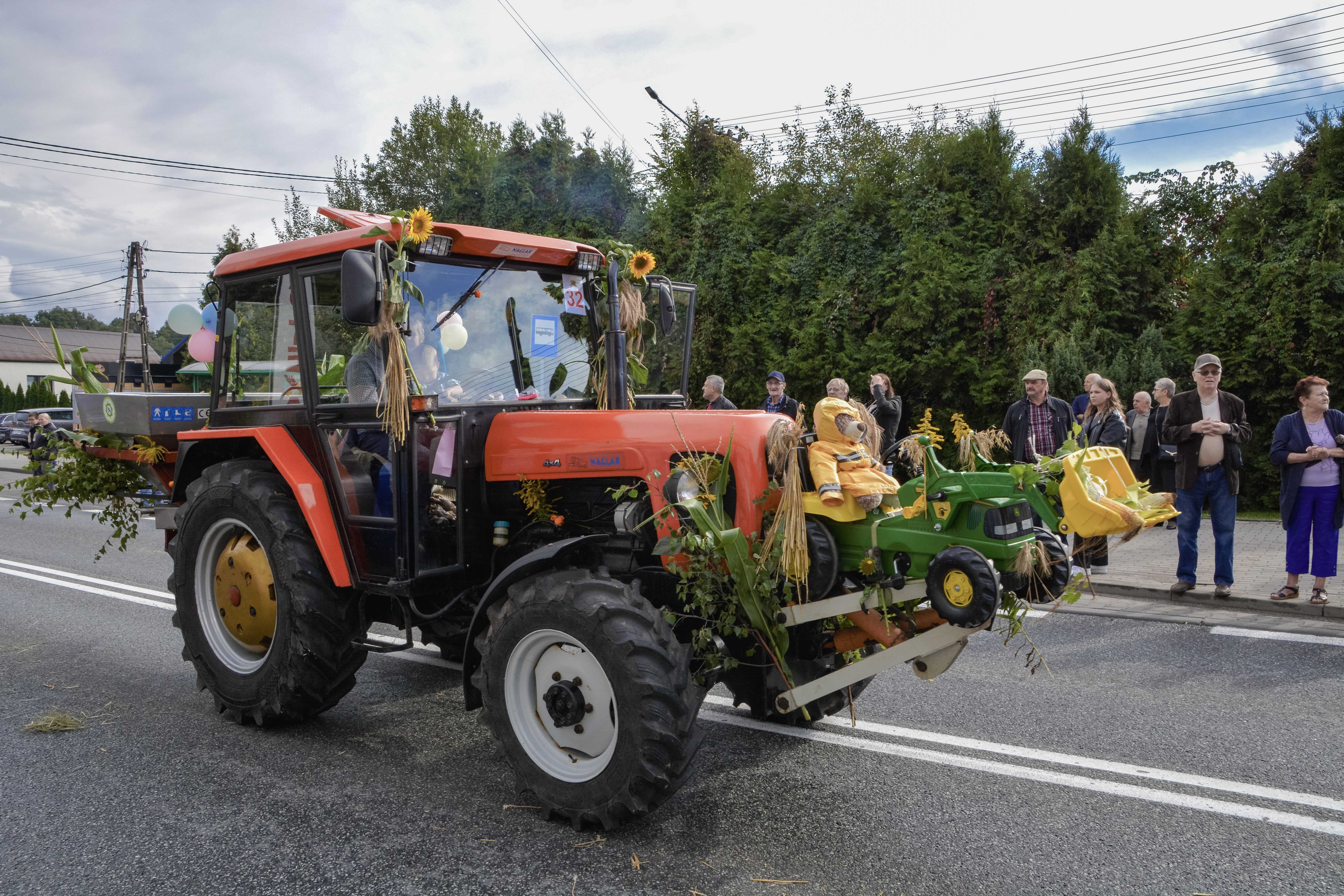 Czerwony traktor przyozdobiony na&nbsp;dożynki w&nbsp;słoneczniki, gałązki brzozy, liście kukurydzy i&nbsp;snopki zboża, wiezie przed sobą mały zabawkowy traktorek dziecięcy z&nbsp;misiem w&nbsp;ż&oacute;łtej pelerynie, na&nbsp;chodniku grupa obserwujących korow&oacute;d os&oacute;b