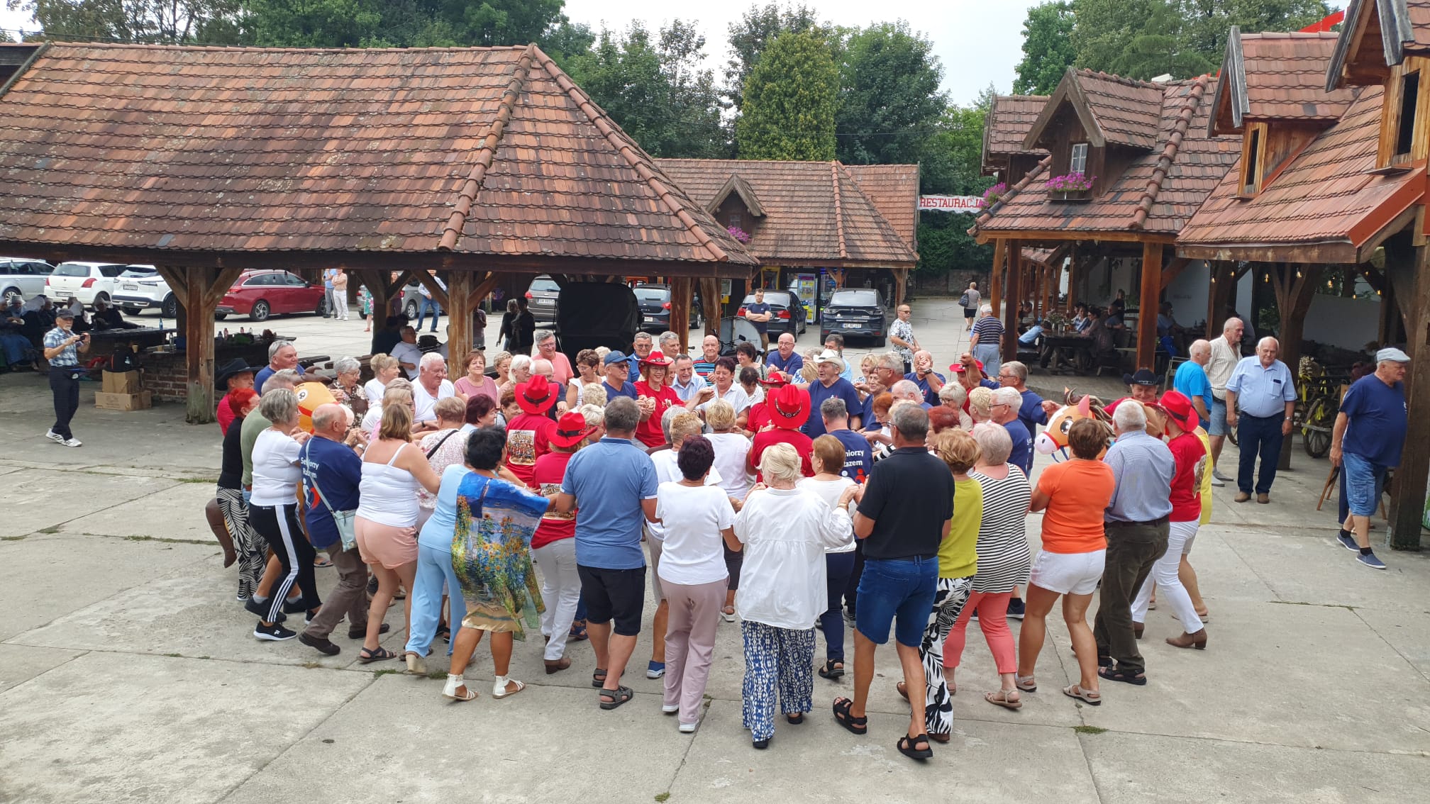 Grupa senior&oacute;w stoi w&nbsp;dużym kole, zwr&oacute;ceni ku sobie, stoją na&nbsp;betonowym placu, wok&oacute;ł zabudowania