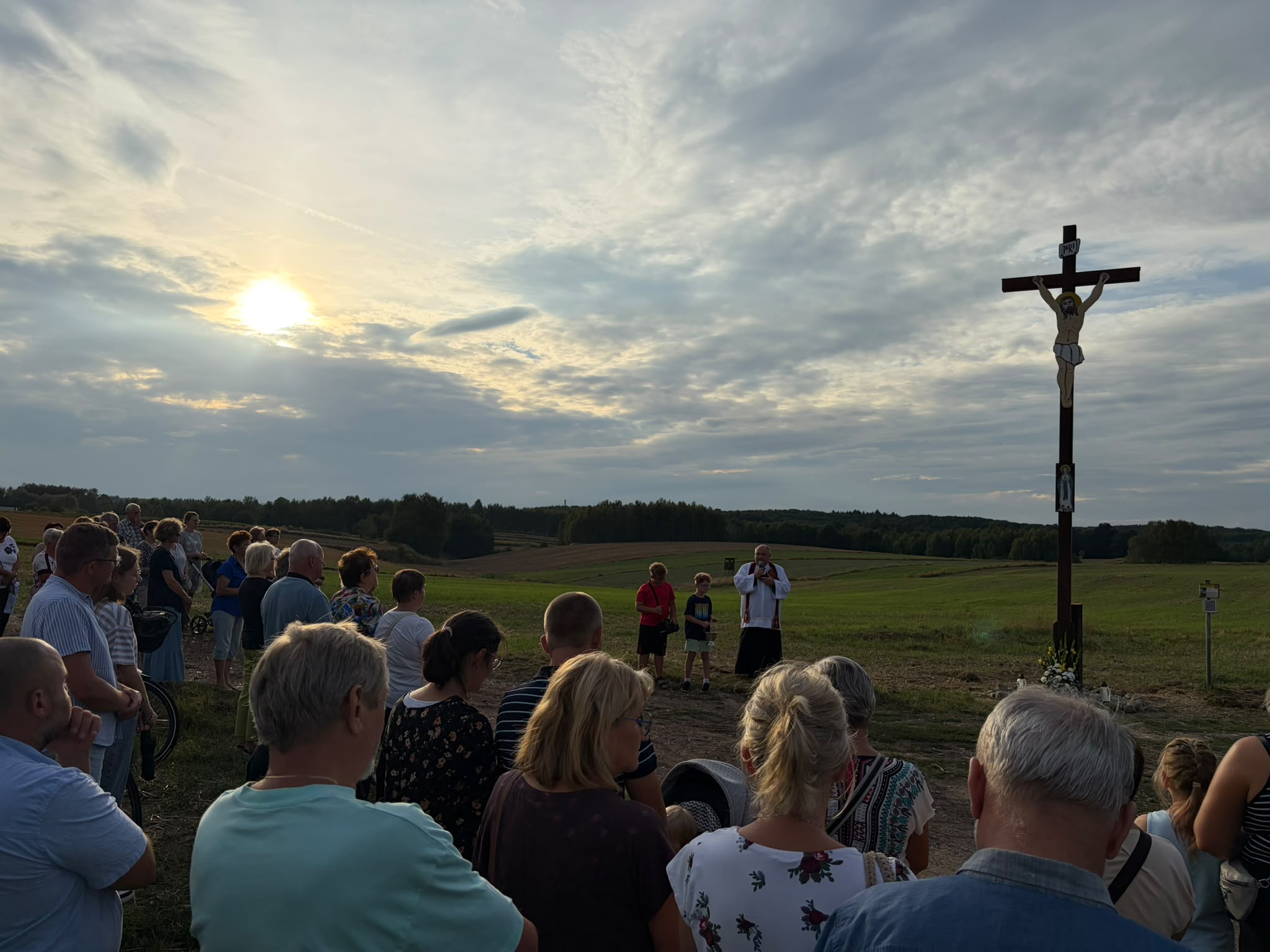 Grupa os&oacute;b stoi przy rozdrożu polnych dr&oacute;g w&nbsp;pobliżu duzego, drewnianego krzyża z&nbsp;wizerunkiem Jezusa, obok stoi ksiądz