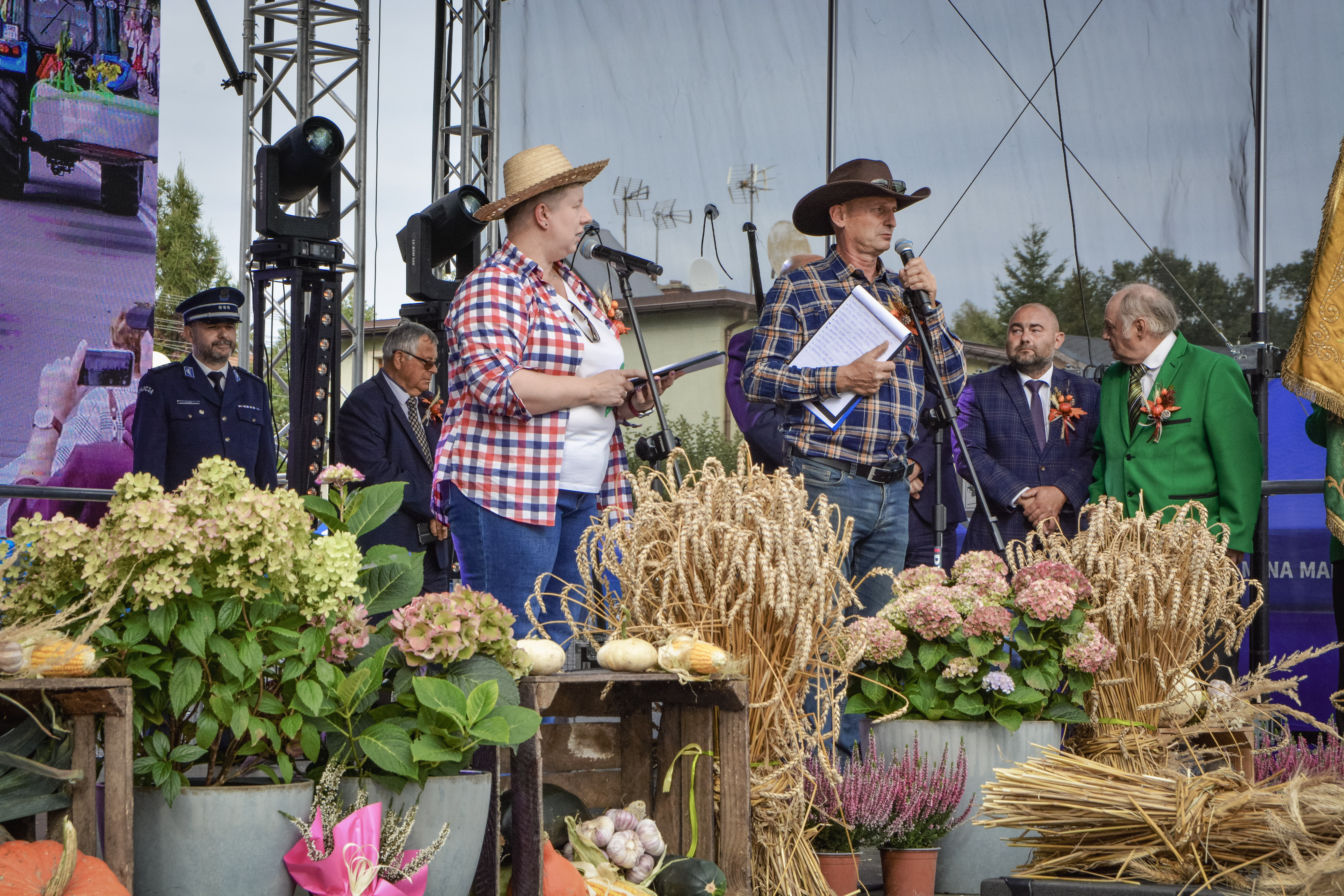 Kobieta i&nbsp;mężczyzna w&nbsp;kraciastych koszulach i&nbsp;słomkowych kapeluszach-prowadzący tegoroczne Dożynki Gminne w&nbsp;Marklowicach, stoją na&nbsp;scenie, w&nbsp;tle zaproszeni goście i&nbsp;władze gminy