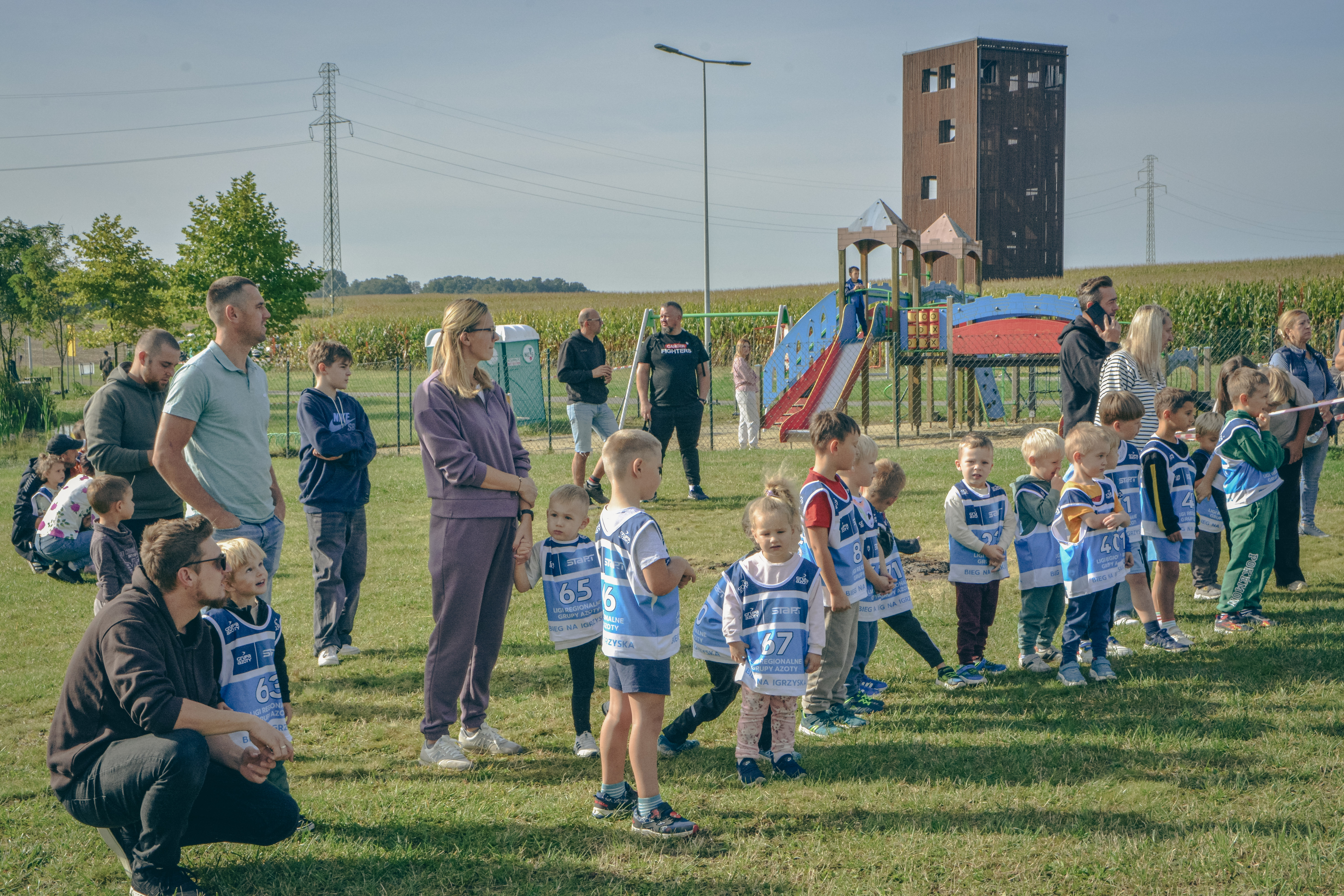 Dzieci wraz z&nbsp;rodzicami na&nbsp;linii startu 27. Otwartego Biegu Ulicznego o&nbsp;Puchar W&oacute;jta Gminy Marklowice, wszyscy stoją na&nbsp;łące na&nbsp;Żr&oacute;dlanej Oazie, w&nbsp;tle wież awidokowa i&nbsp;pole kukurydzy