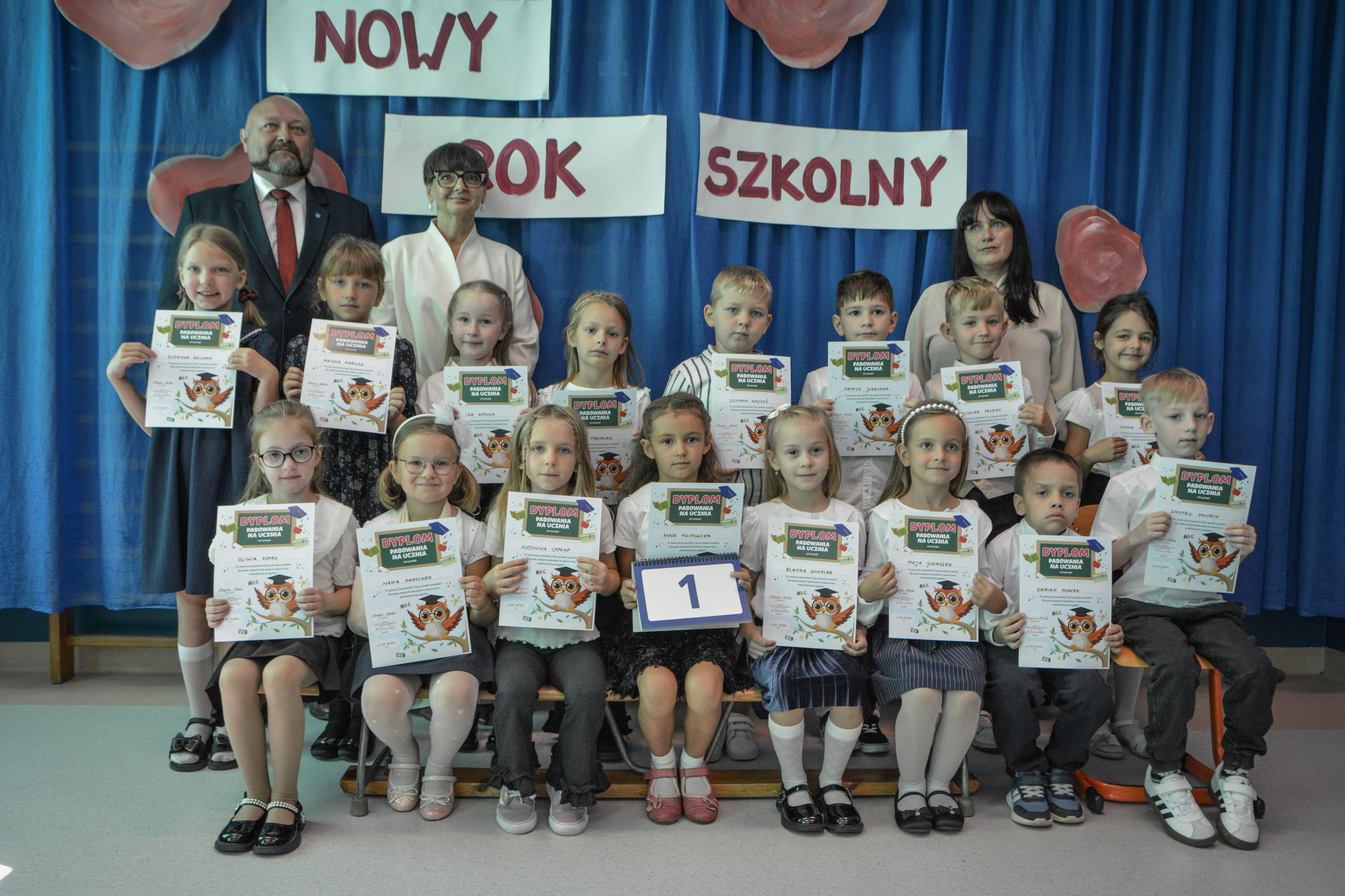 Chłopcy i&nbsp;dziewczynki w&nbsp;odświętnych strojach stoją oraz&nbsp;siedzą z&nbsp;dyplomami podczas uroczystej inauguracji nowego roku szkolnego