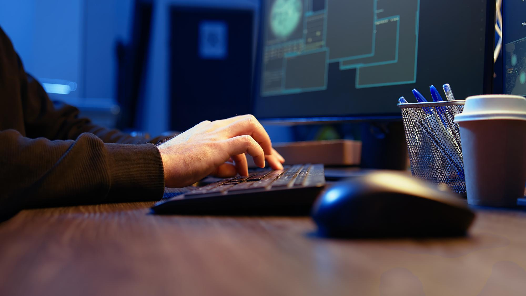 W kadrze ręce siedzącej przy biurku postaci, na&nbsp;biurku monitor, mysz komputerowa oraz&nbsp;klawiatura, dłonie postaci znajdują się nad klawiaturą, na&nbsp;monitorze ciemny ekran i&nbsp;tabelki