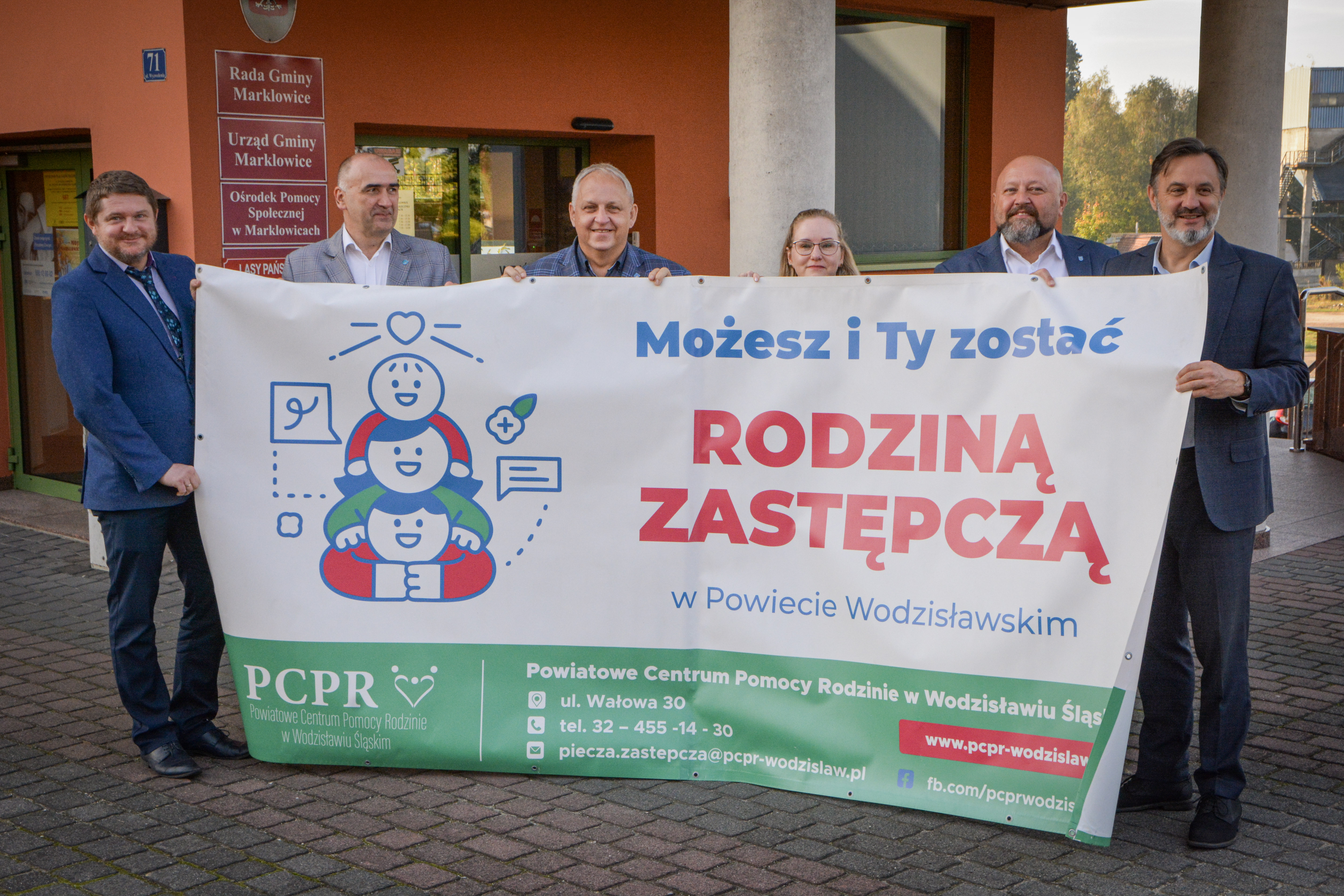 Kilka os&oacute;b stoi przed budynkiem Urzędu Gminy w&nbsp;Marklowicach, trzymając rozwinięty duży baner promocyjny o&nbsp;rodzinach zastępczych