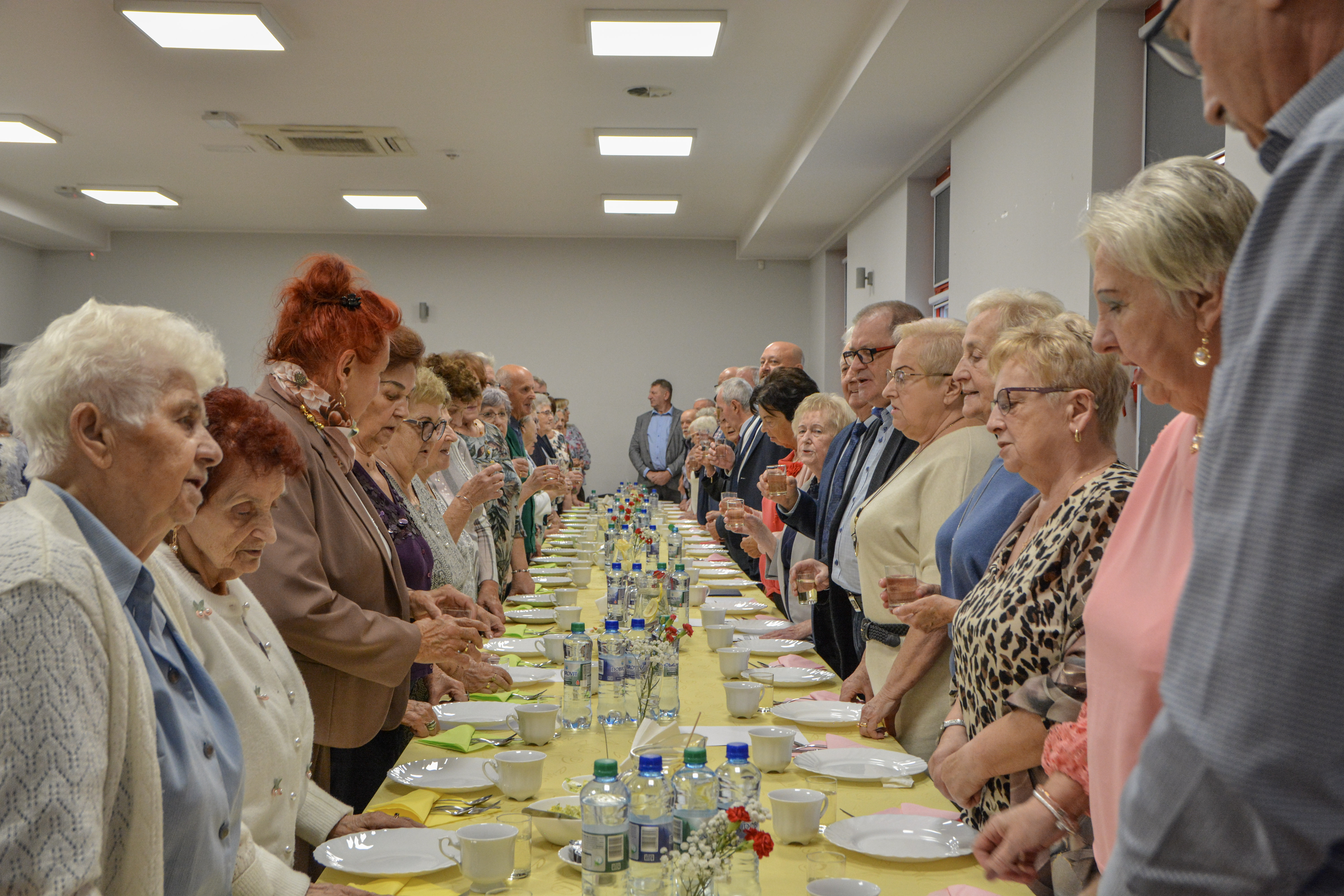Seniorzy stoją przy zastawionym stole, wsp&oacute;lnie śpiewając "sto lat"