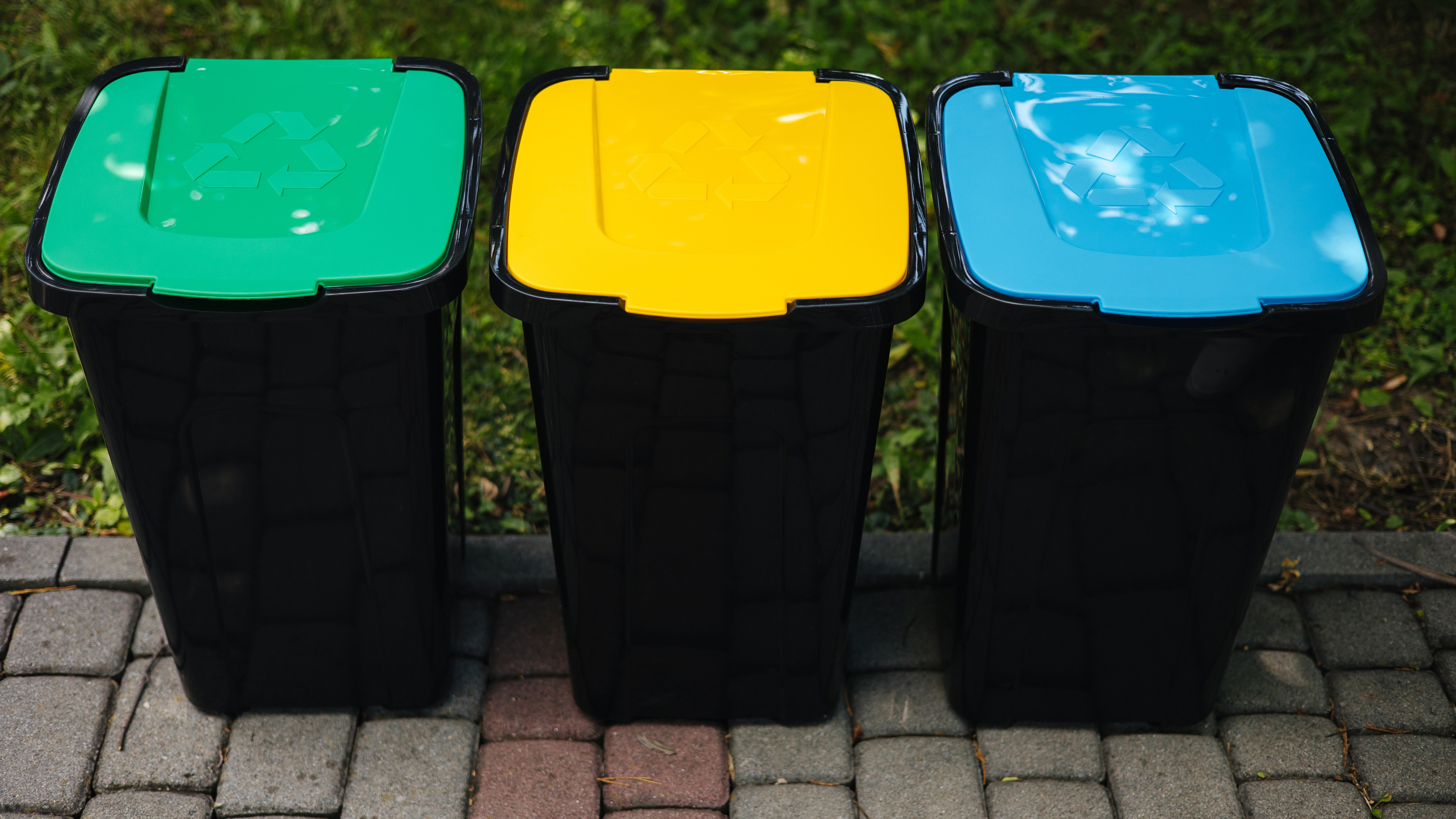 Trzy czarne pojemniki na&nbsp;śmieci z&nbsp;kolorowymi pokrywkami stoją na&nbsp;podjeździe