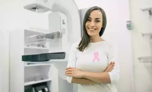 Pani w białej koszulce z różową wstążeczką na piersi pozuje przed aparatem do badania mammograficznego