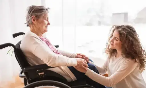 Starsza pani na wózku inwalidzkim z uśmiechem trzyma ręce swojej opiekunki, w tle za oknem zimowy krajobraz