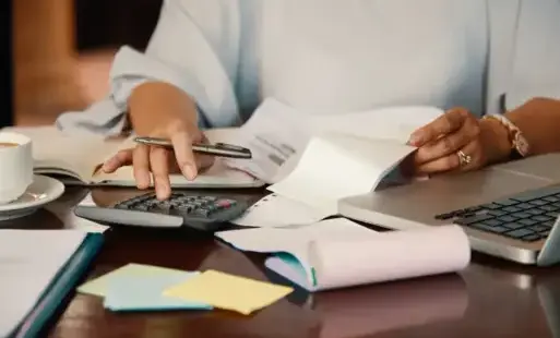 Stół z porozkładanymi dokumentami i rachunkami, na stole kalkulator oraz laptop, czyjeś dłonie trzymają w jednej ręce długopis, w drugiej rachunek