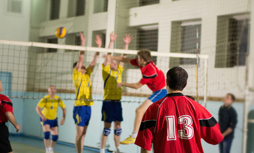 Kilka osób na sali sportowej podzielona na dwie drużyny, jedni w żółtych drudzy w czerwonych koszulkach, grają w siatkówę