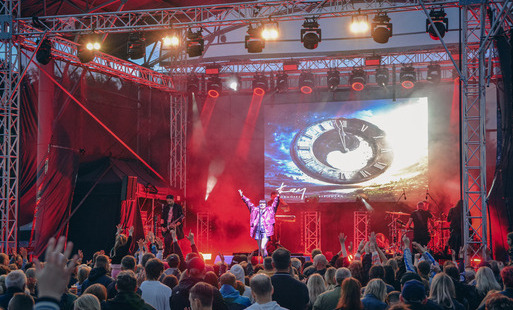 Widoczna scena z czerwonymi światłami, na scenie stoi postać z rękoma uniesionymi w górze, przed sceną tłum fanów