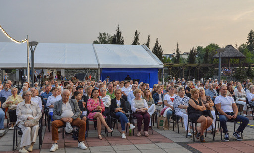 Duża grupa ludzi siedzi na krzesłach na dużym placu, w tle namiot eventowy, obok świetlna girlanda zawieszona na słupku