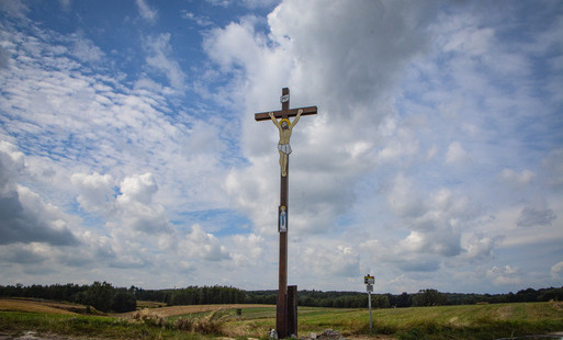 Polne rozdroże, przy nim stoi duży drewniany krzyż z wizerunkiem umęczonego Jezusa, pod spodem mniejszy wizerunek Matki Boskiej, w tle pola uprawne i lasy