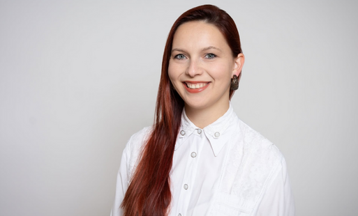 Młoda, uśmiechnięta kobieta pozuje do zdjęcia, w białej koszuli, na jednolitym szarym tle