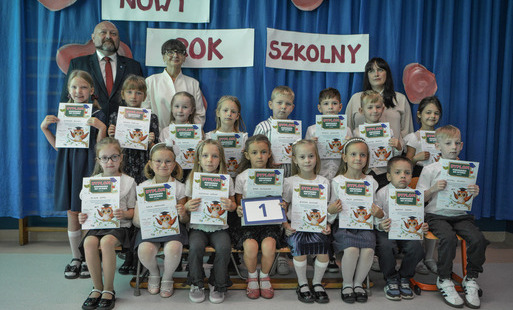 Chłopcy i dziewczynki w odświętnych strojach stoją oraz siedzą z dyplomami podczas uroczystej inauguracji nowego roku szkolnego