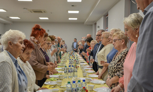 Seniorzy stoją przy zastawionym stole, wspólnie śpiewając "sto lat"